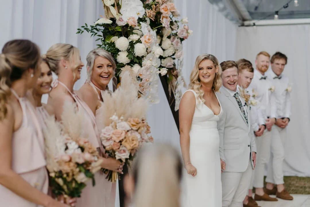 Bride and bridesmaids smiling beside the floral ceremony arch