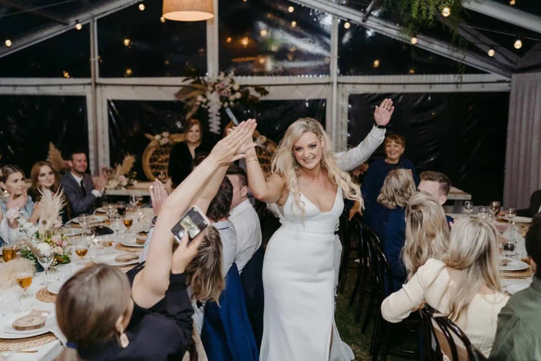 Bride walking between dining tables as seated guests raise their hands