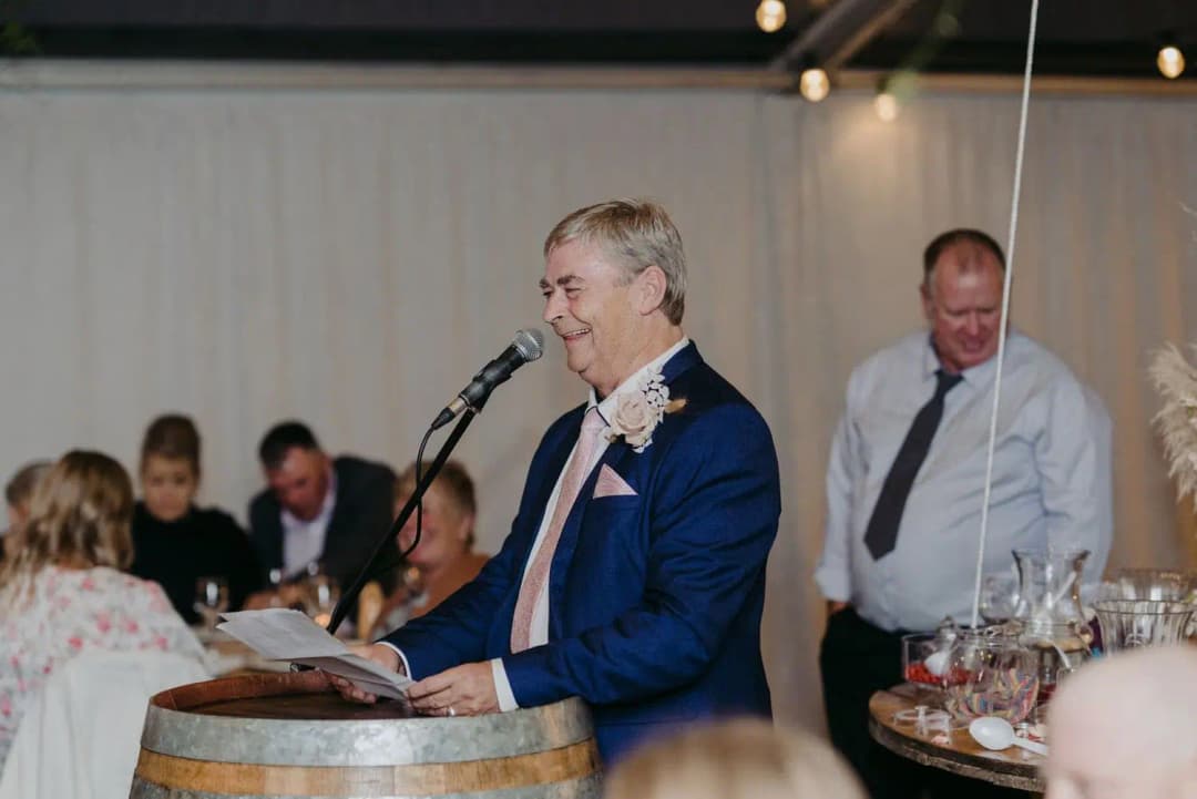 Older man reading a speech into a microphone beside a wooden barrel