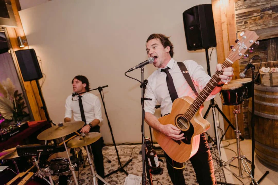 Singer with an acoustic guitar performing beside a drummer indoors