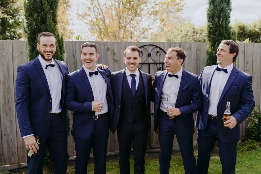 Five men in navy suits smiling together in a garden with drinks