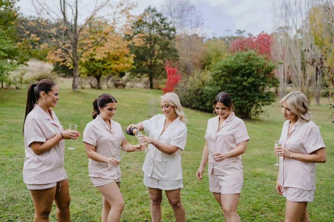 Women in pale pink sleepwear gathering on a lawn with glasses and a bottle