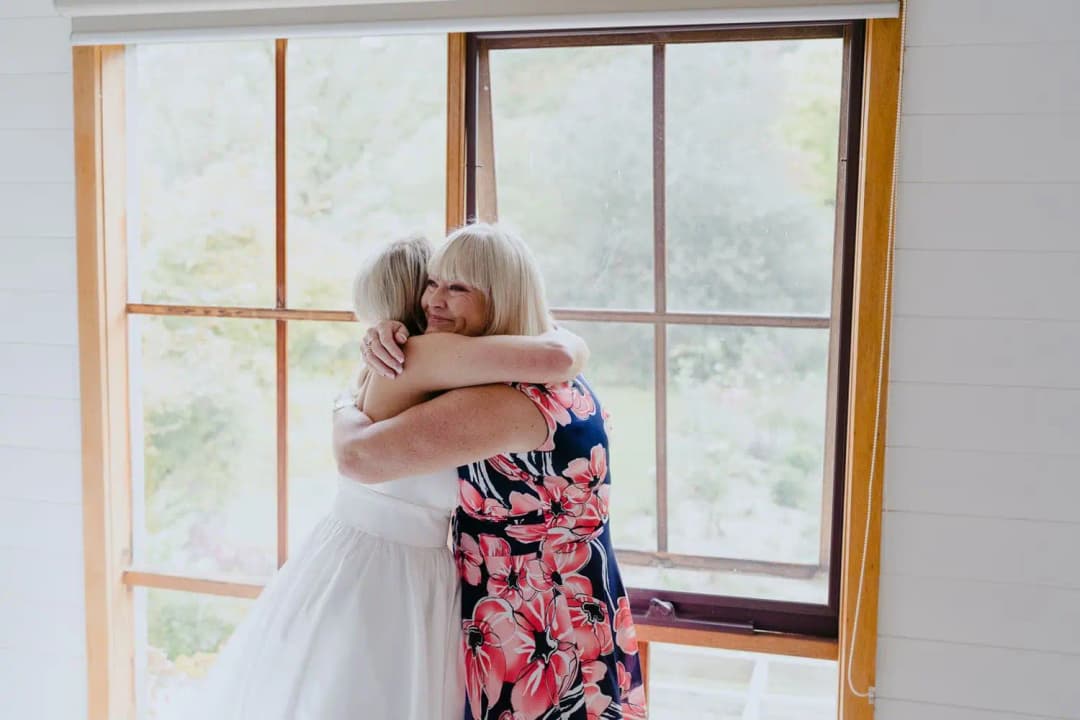 Bride embracing a woman in a floral dress beside a window