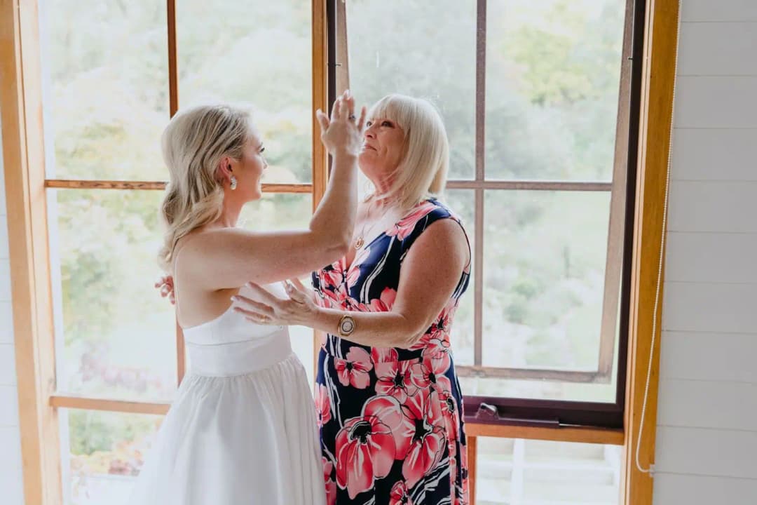 Bride touching a woman's face while they smile beside a window