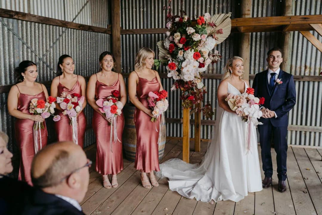 Bride and groom standing together beside four bridesmaids beneath a floral arch