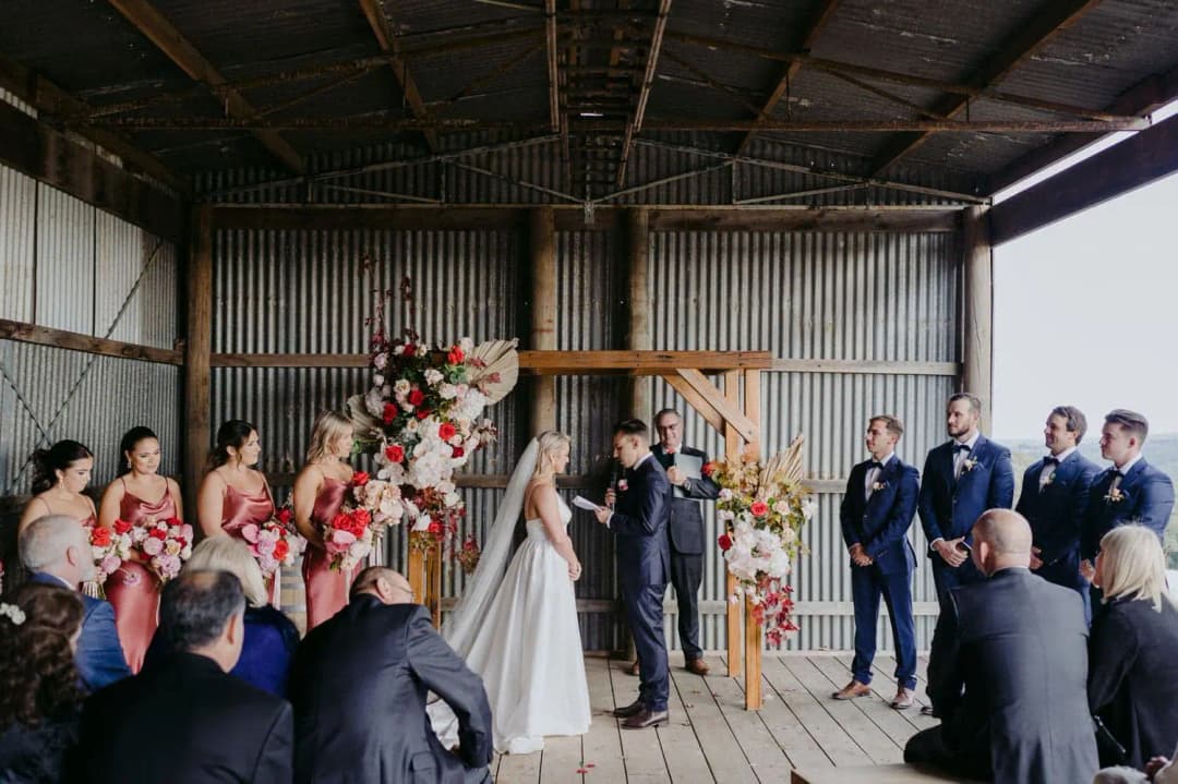Wide view of the couple and wedding party beneath a floral arch inside the shed