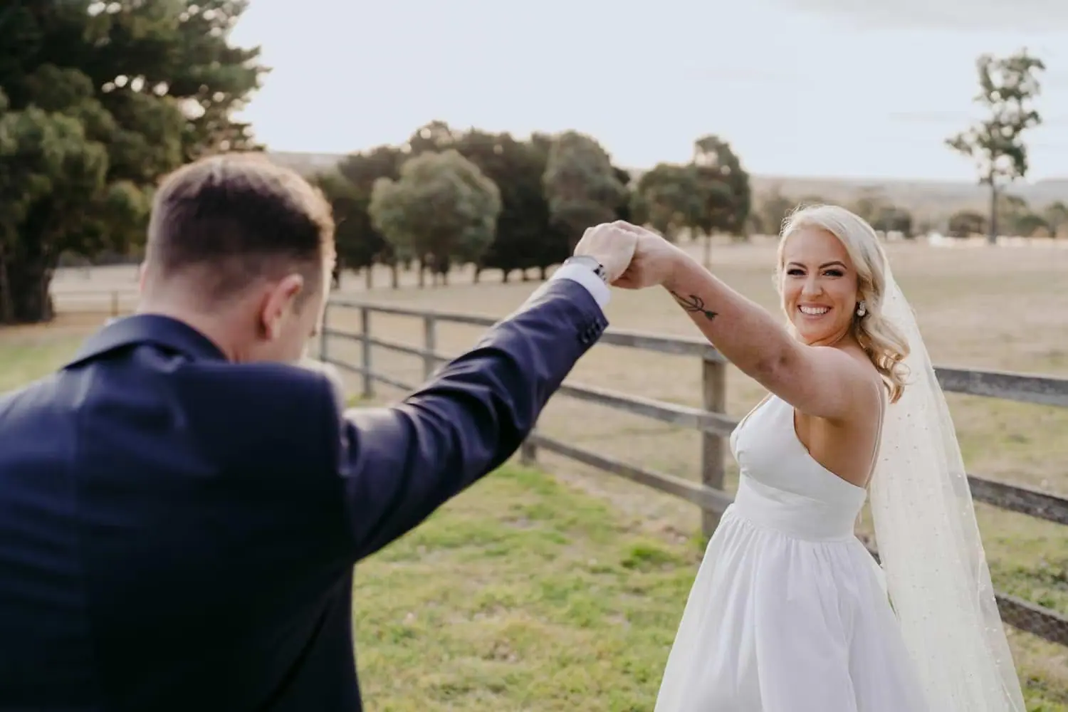 Groom holding the bride's hand above her head beside a timber fence