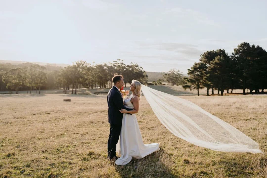 Bride and groom embracing in a sunlit field with her veil blowing behind them