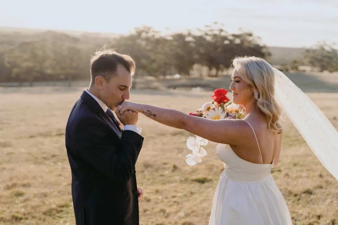 Groom kissing the bride's hand while she holds her bouquet in a field