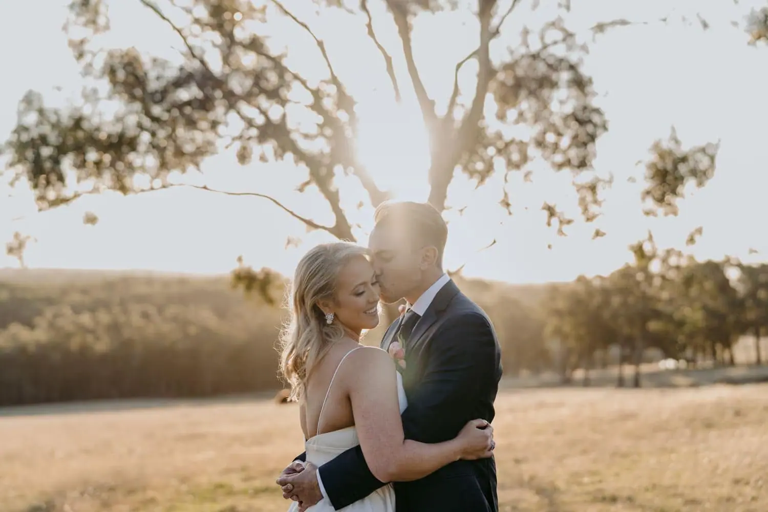 Bride and groom embracing beneath a tree in warm evening sunlight