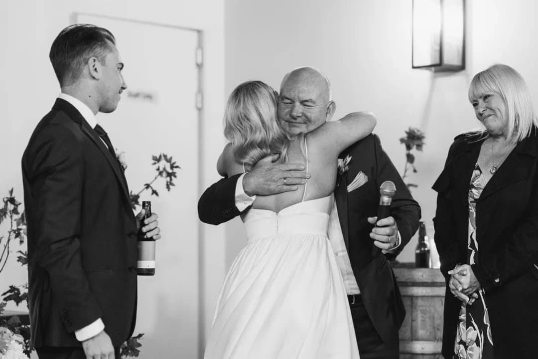 Black and white photograph of the bride hugging a bald man beside the groom