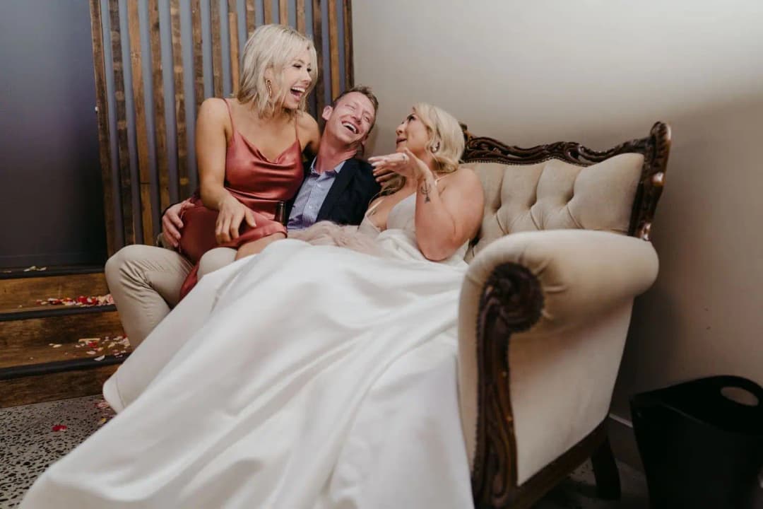 Bride and a bridesmaid laughing with a guest on a pale sofa