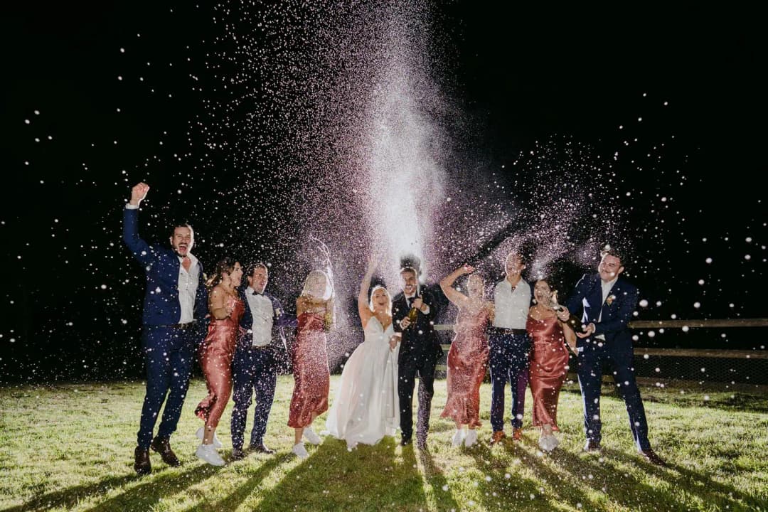 Wedding party cheering beneath a spray of sparkling wine outdoors at night