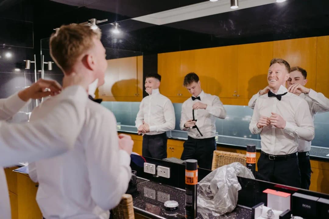 Groom and groomsmen adjusting their bow ties in front of a mirror