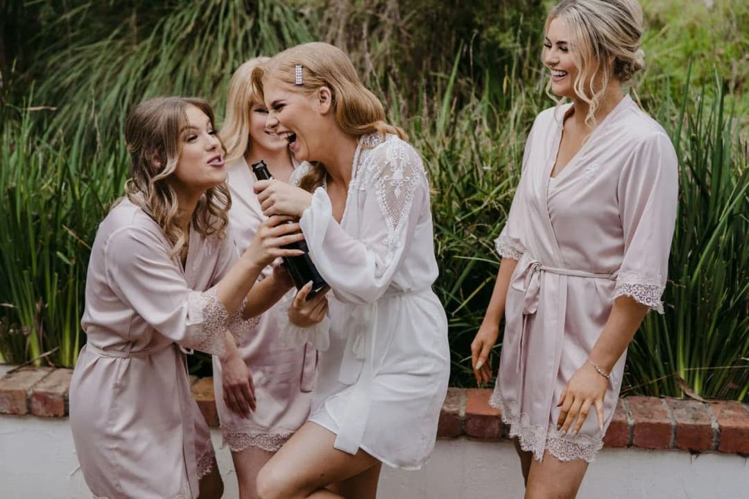Women in pale robes laughing around the bride with a drink