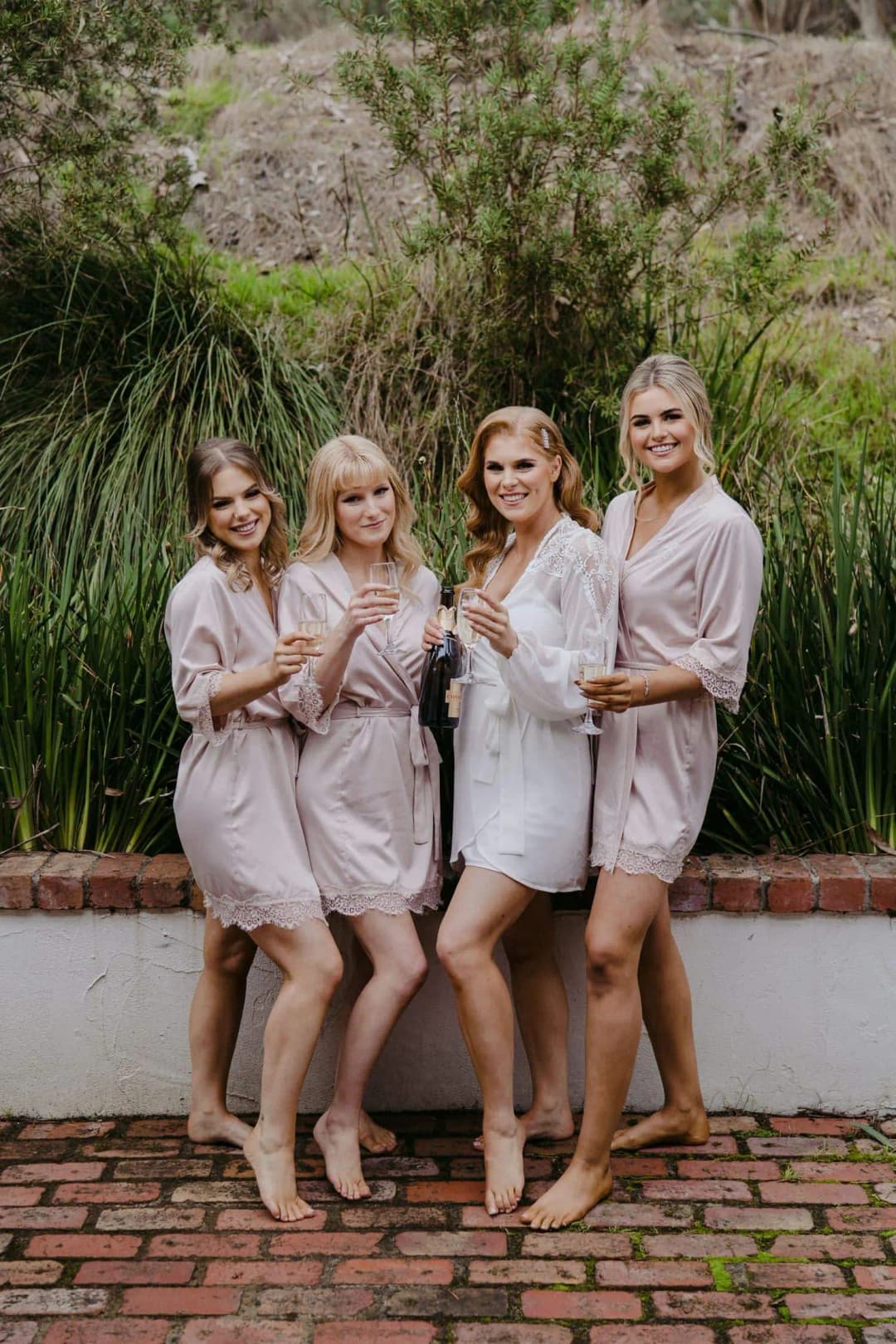 Bride and bridesmaids in pale robes holding glasses in a garden