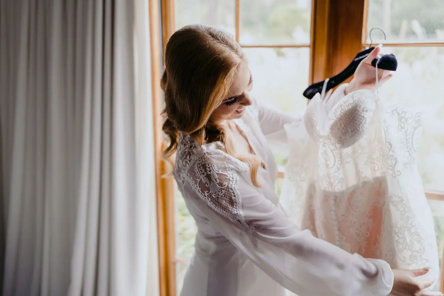 Bride looking at her wedding dress hanging beside a window