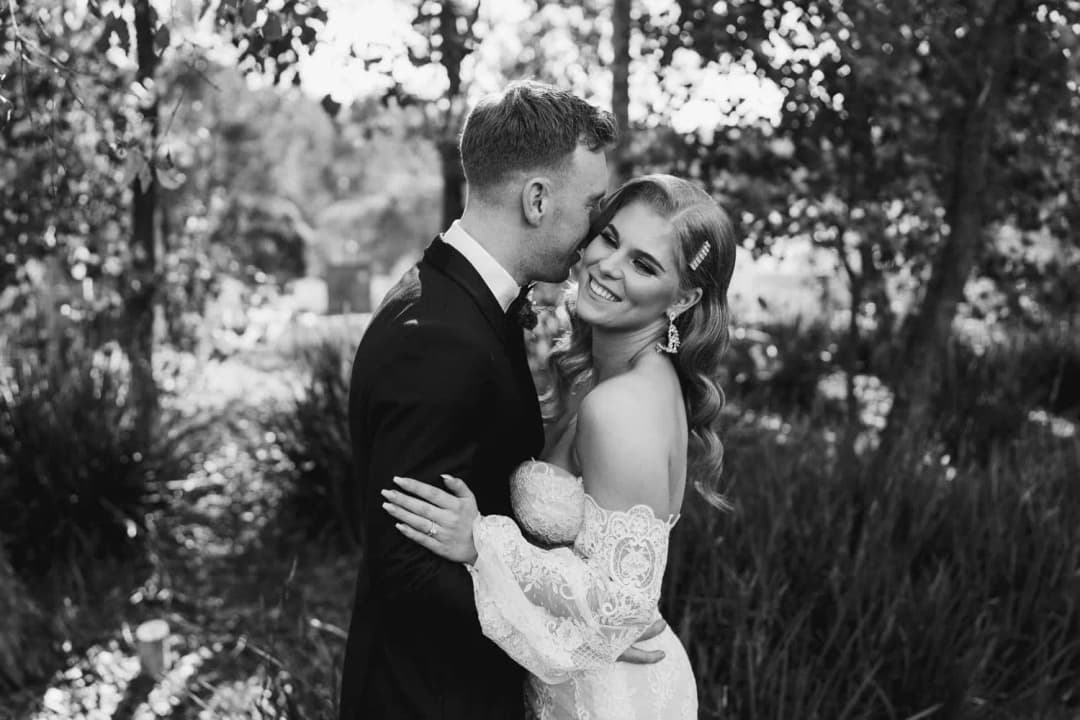 Black-and-white photograph of the groom kissing the bride beside garden trees