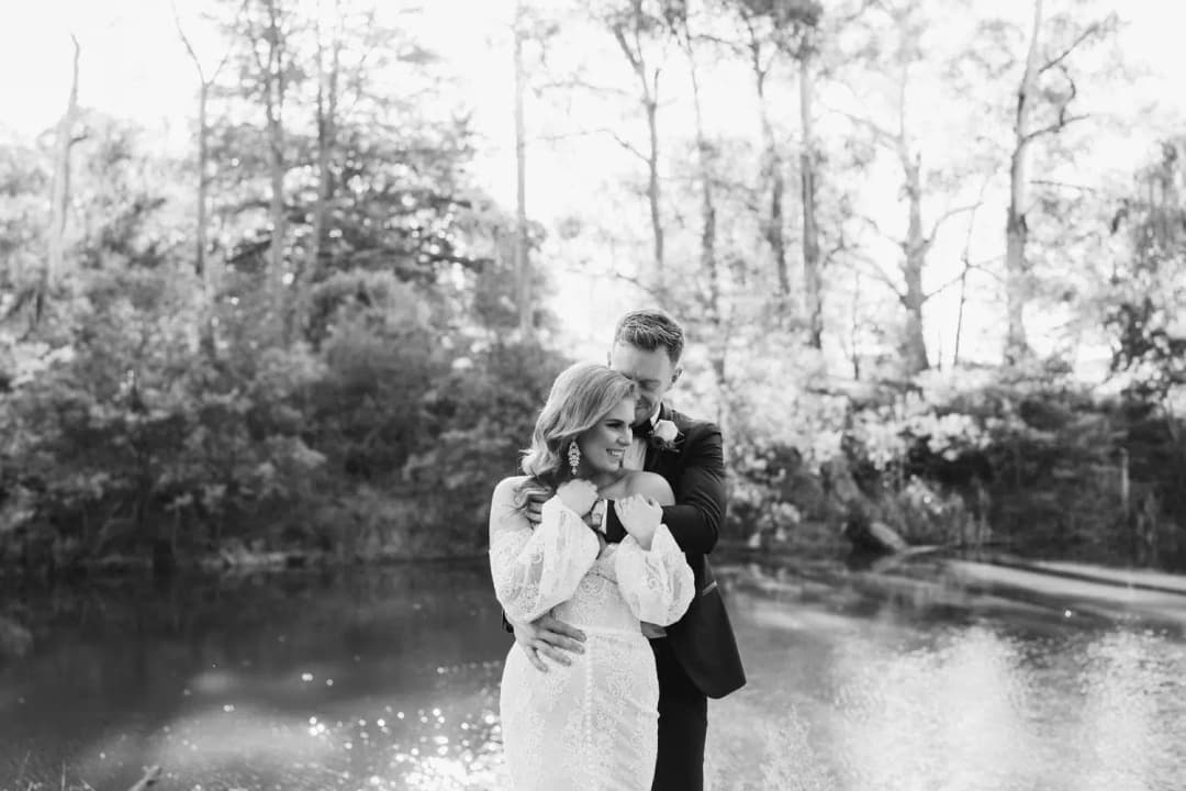 Black-and-white photograph of the groom embracing the bride beside a pond
