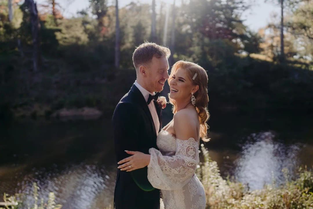 Bride and groom smiling at each other beside water under tall trees