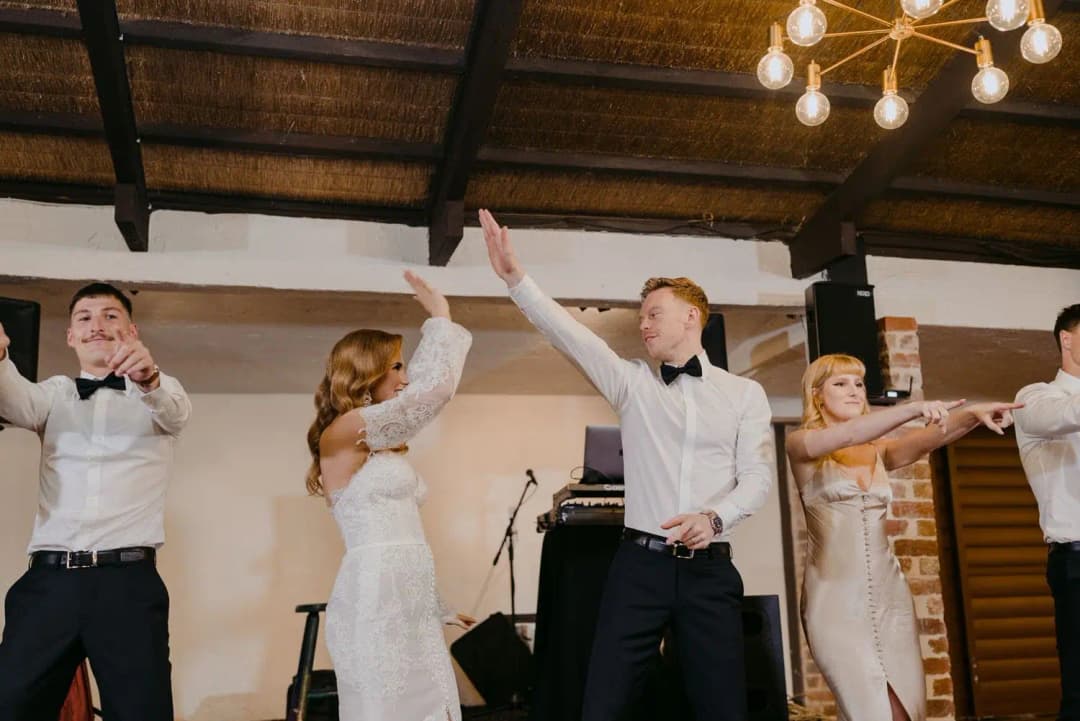Bride and groomsmen dancing with raised arms beneath a chandelier