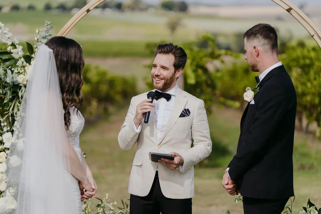 Celebrant speaking into a microphone beneath a floral arch beside the couple.