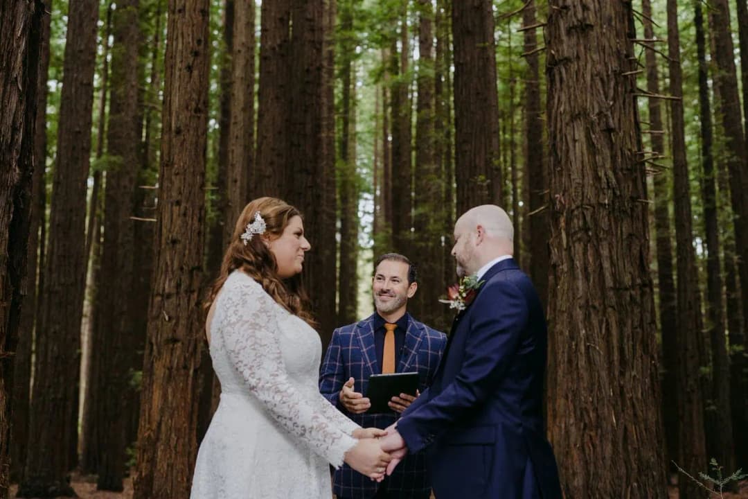 Couple holding hands beside their celebrant among tall forest trees.