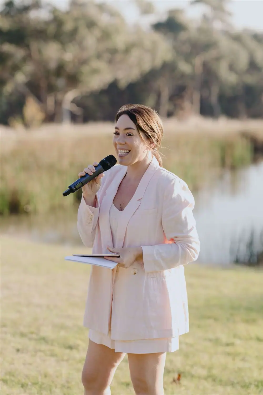 Woman in a pale pink suit speaking into a microphone beside a pond.