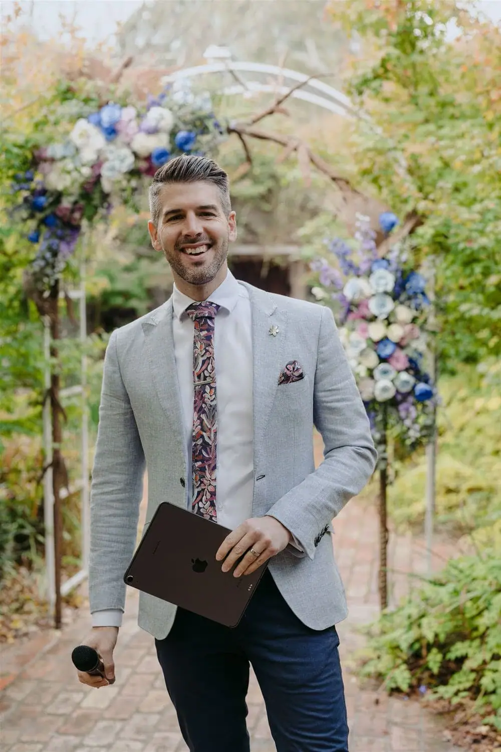 Man holding a microphone and folder beneath a floral garden arch.