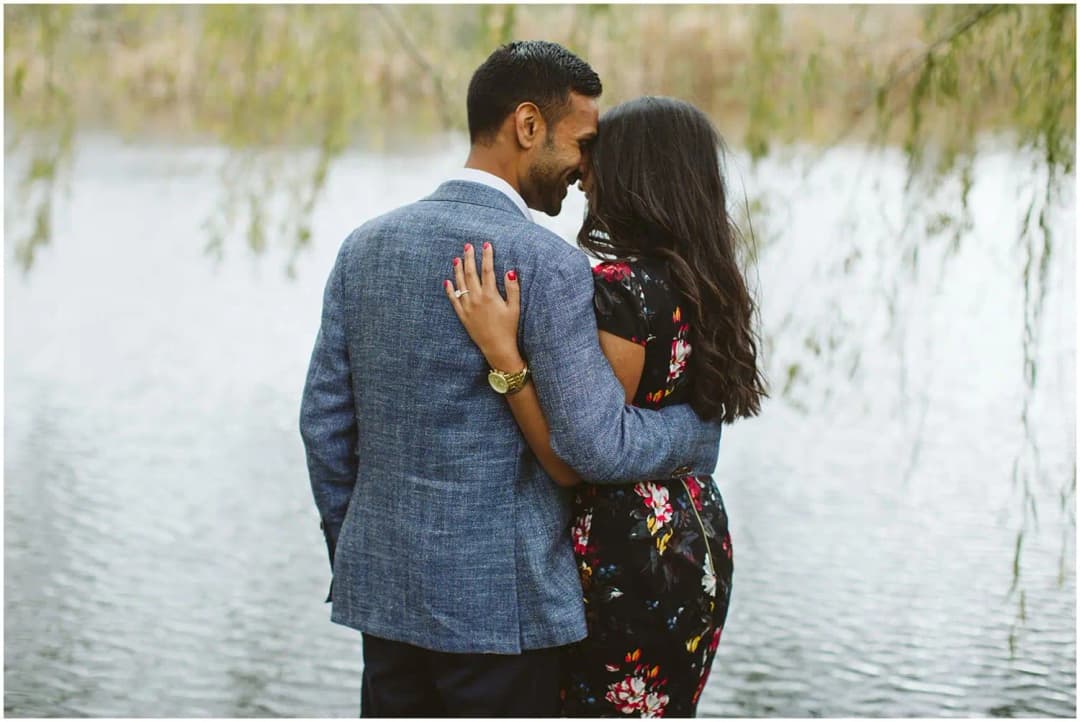 Couple embracing beside water beneath overhanging willow branches.