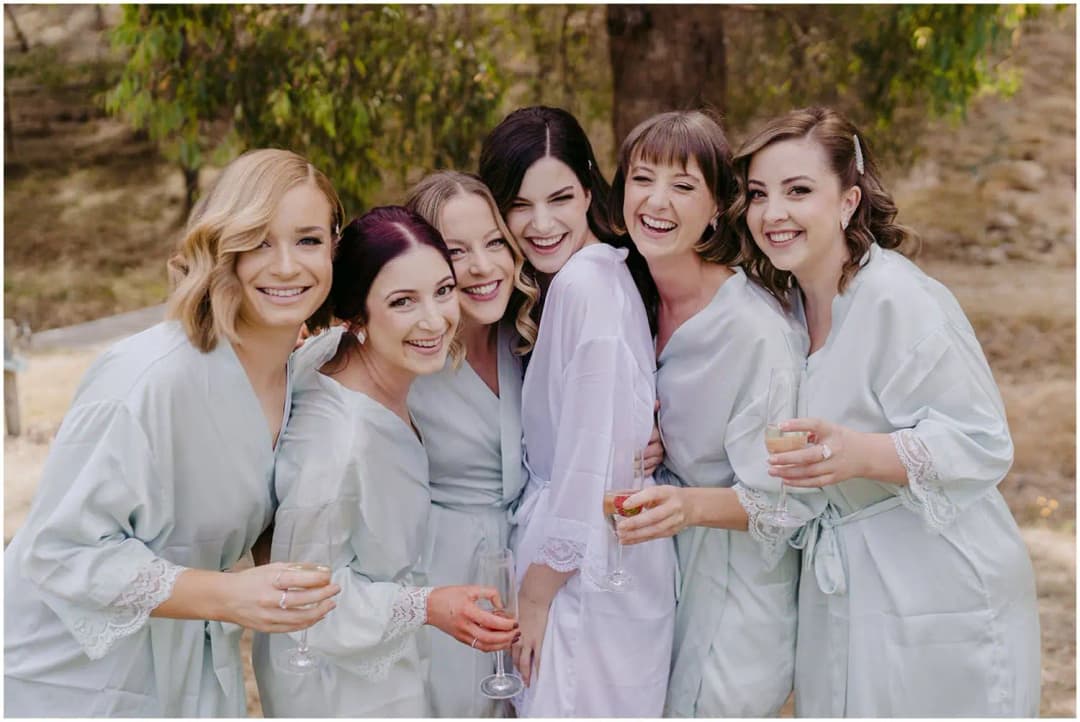 Six women smiling together outdoors in pale robes while holding drinks