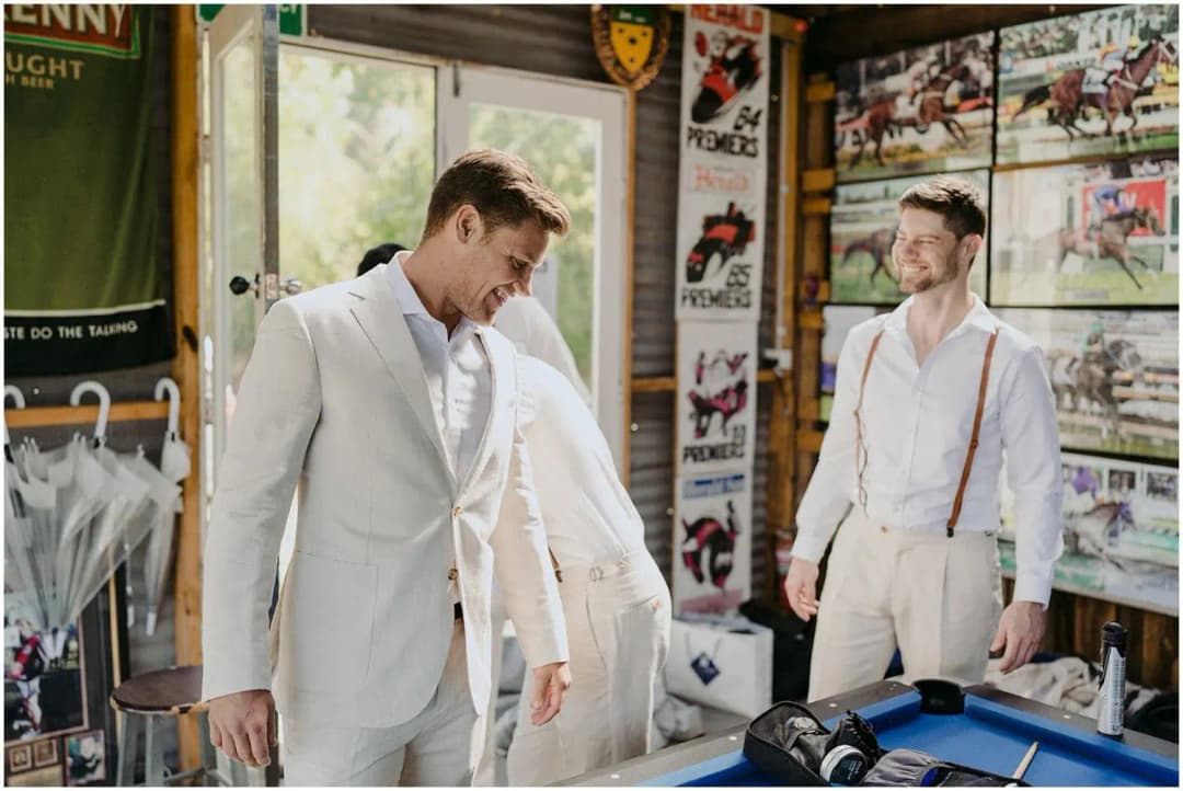 Groom putting on a pale suit jacket while a friend watches beside the pool table