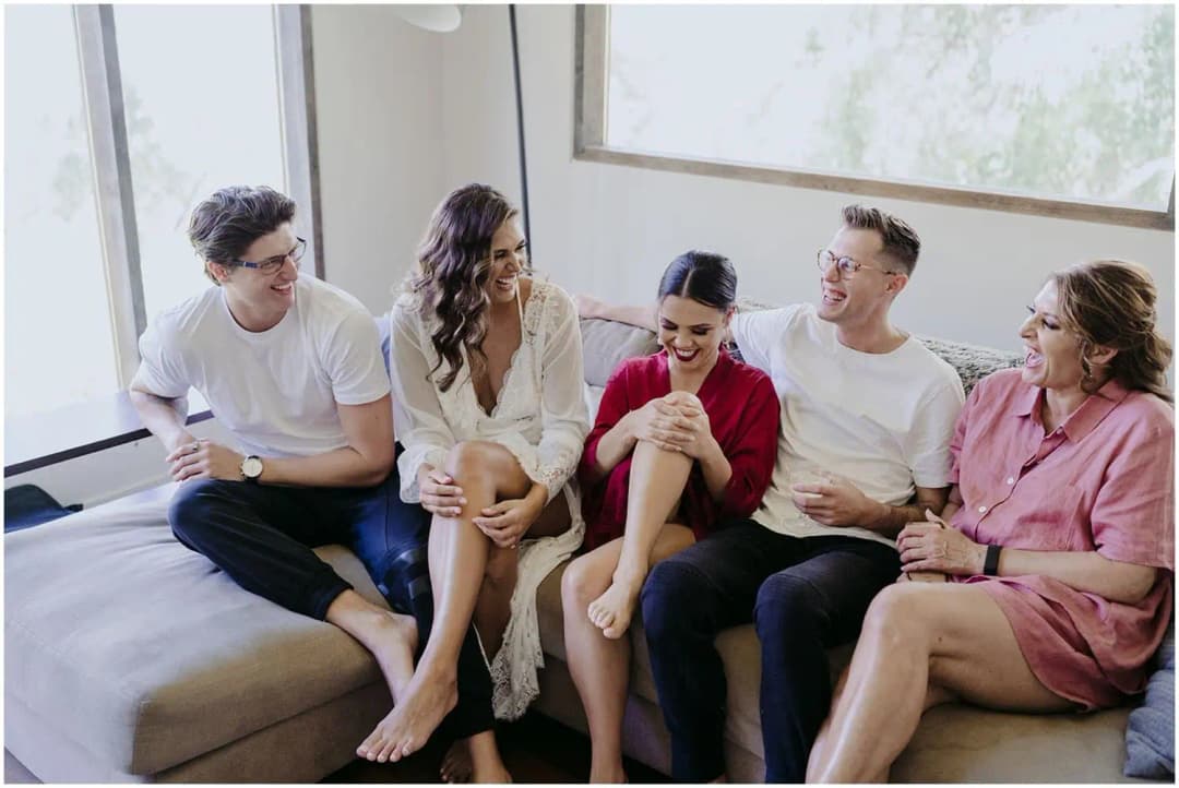 Five friends laughing together while seated on a bed beside a window