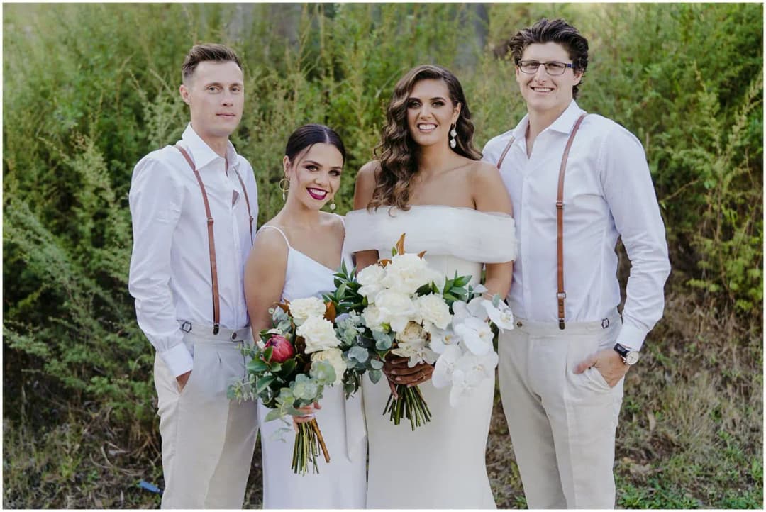 Bride and three friends posing outdoors with white flowers against green hedges