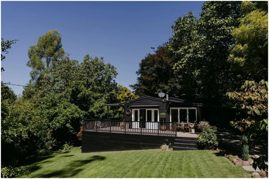 Dark timber cottage with a veranda overlooking a green lawn and garden