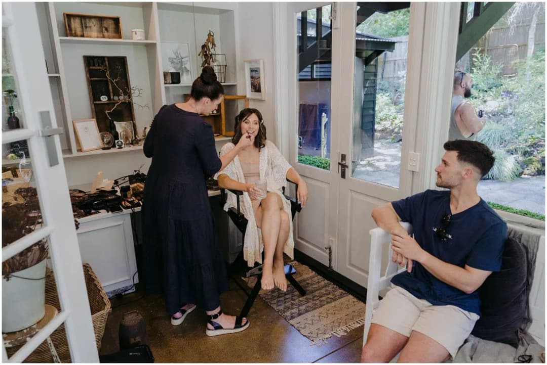 Makeup artist working with a seated bride while a man relaxes with a mug