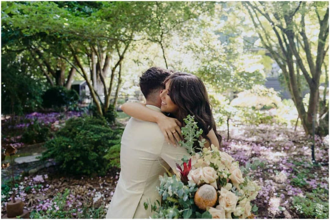 Bride and groom embracing in a leafy garden with a bouquet in the foreground