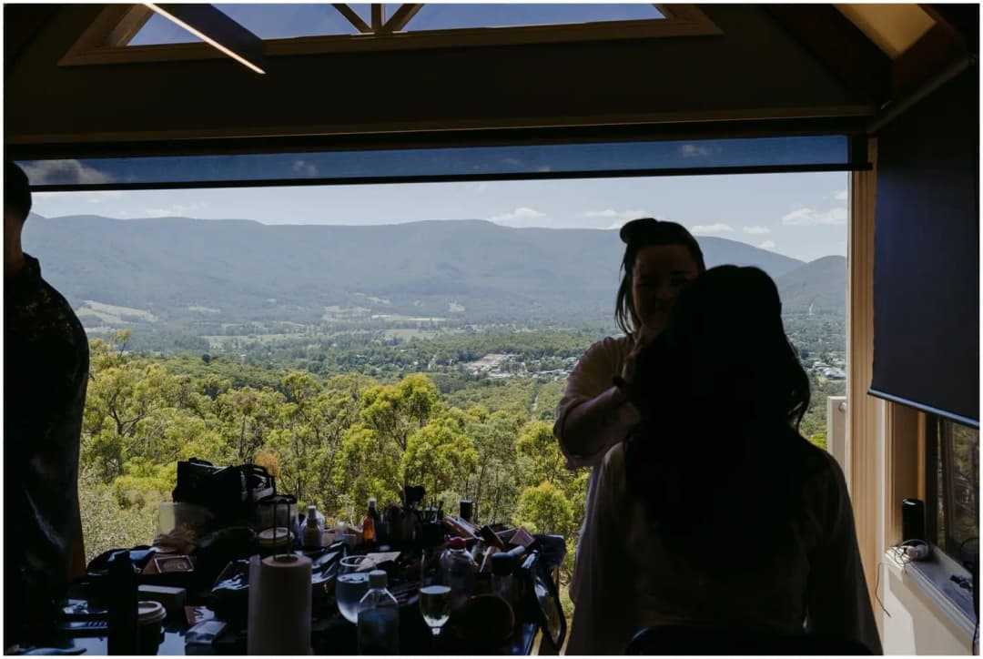 Makeup artist working with a seated woman beside a wide window overlooking a valley