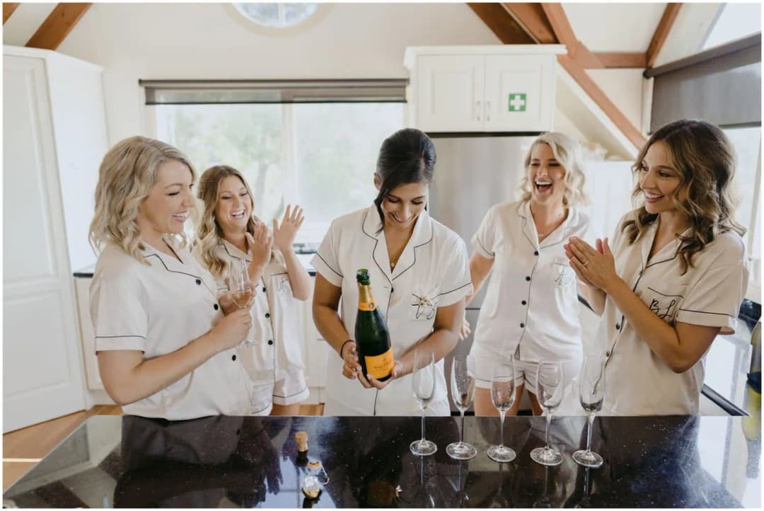 Women laughing around a kitchen counter as one opens a bottle of sparkling wine