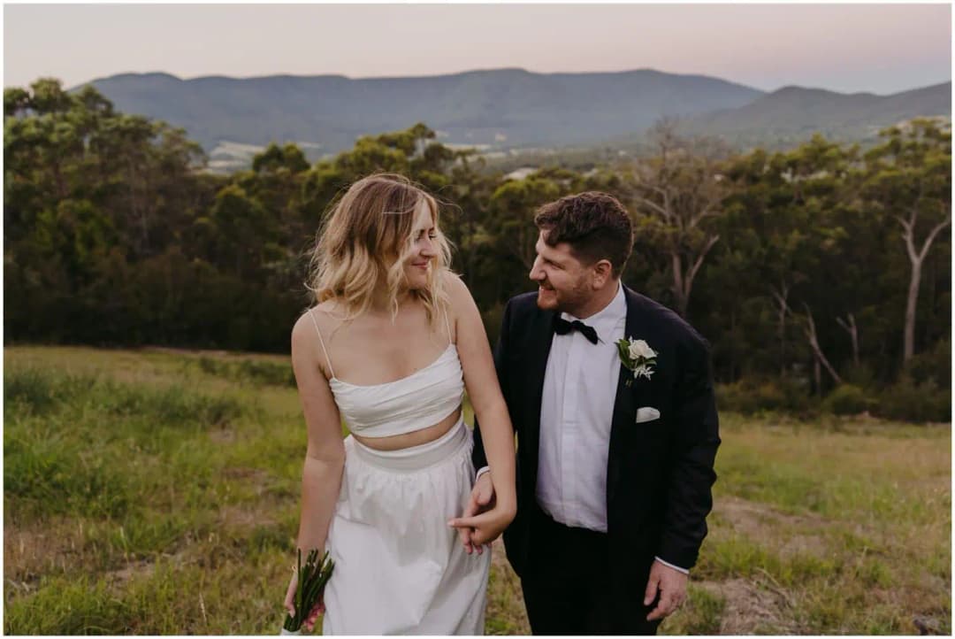 Bride and groom holding hands in a grassy field beneath distant hills