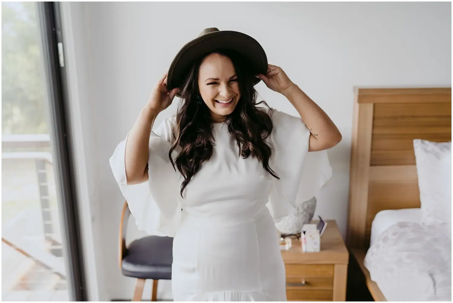 Bride laughing as she adjusts a dark wide-brimmed hat while seated on a bed