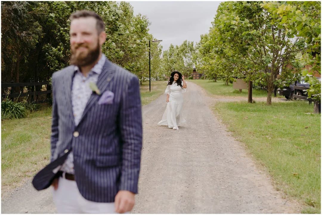 Bride walking along a gravel lane toward the groom beneath leafy trees