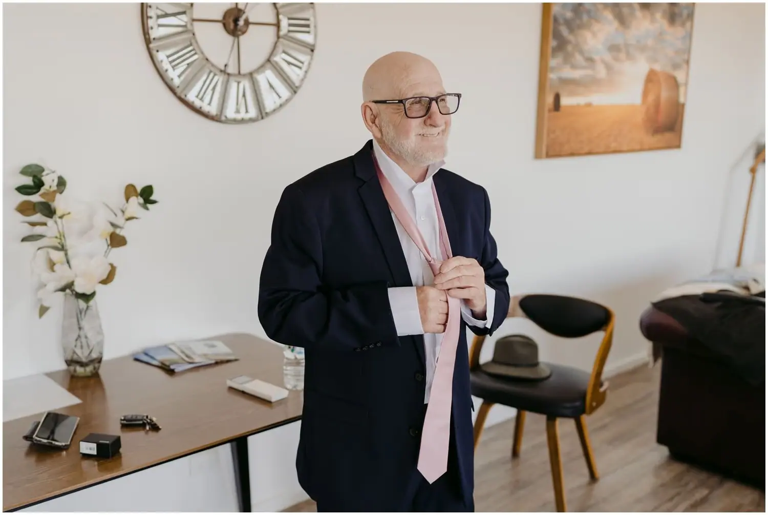 Man in glasses and a dark suit tying a pale pink tie beside a dining table