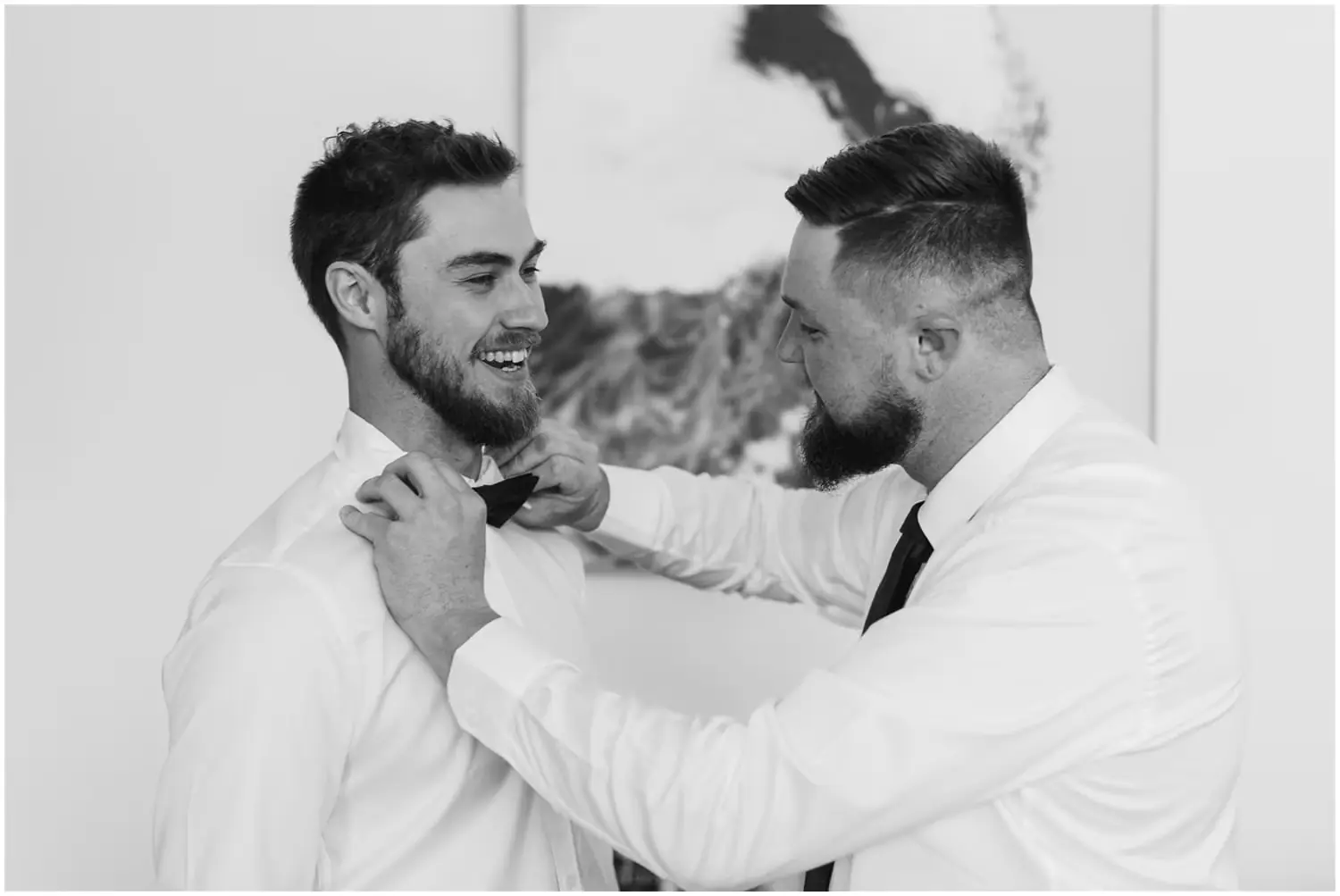 Black and white photograph of a friend adjusting the groom's bow tie
