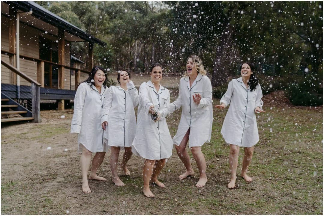 Five women celebrating beneath a spray of sparkling wine beside a timber cabin