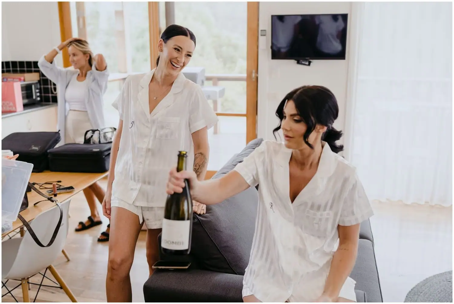 Women in white sleepwear laughing with a bottle of sparkling wine indoors