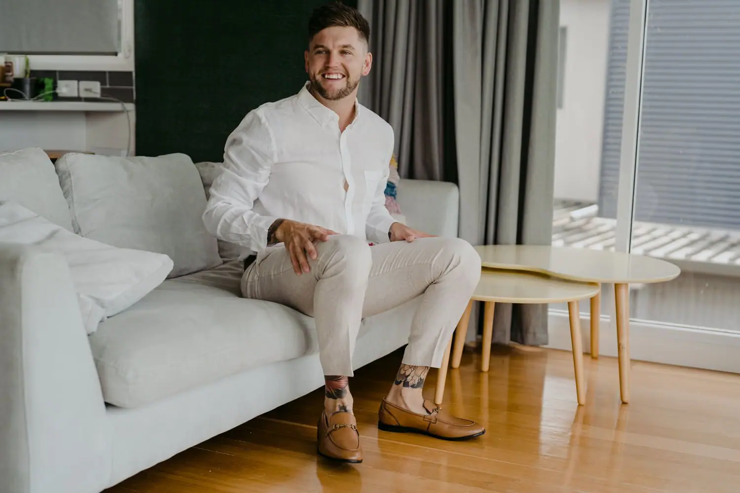 Groom smiling while seated on a white sofa in a shirt and cream trousers