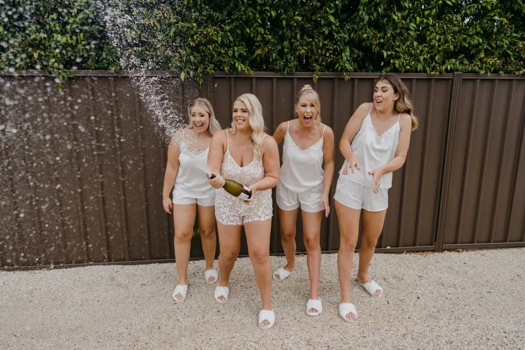 Four women laughing as one sprays a bottle beside a timber fence