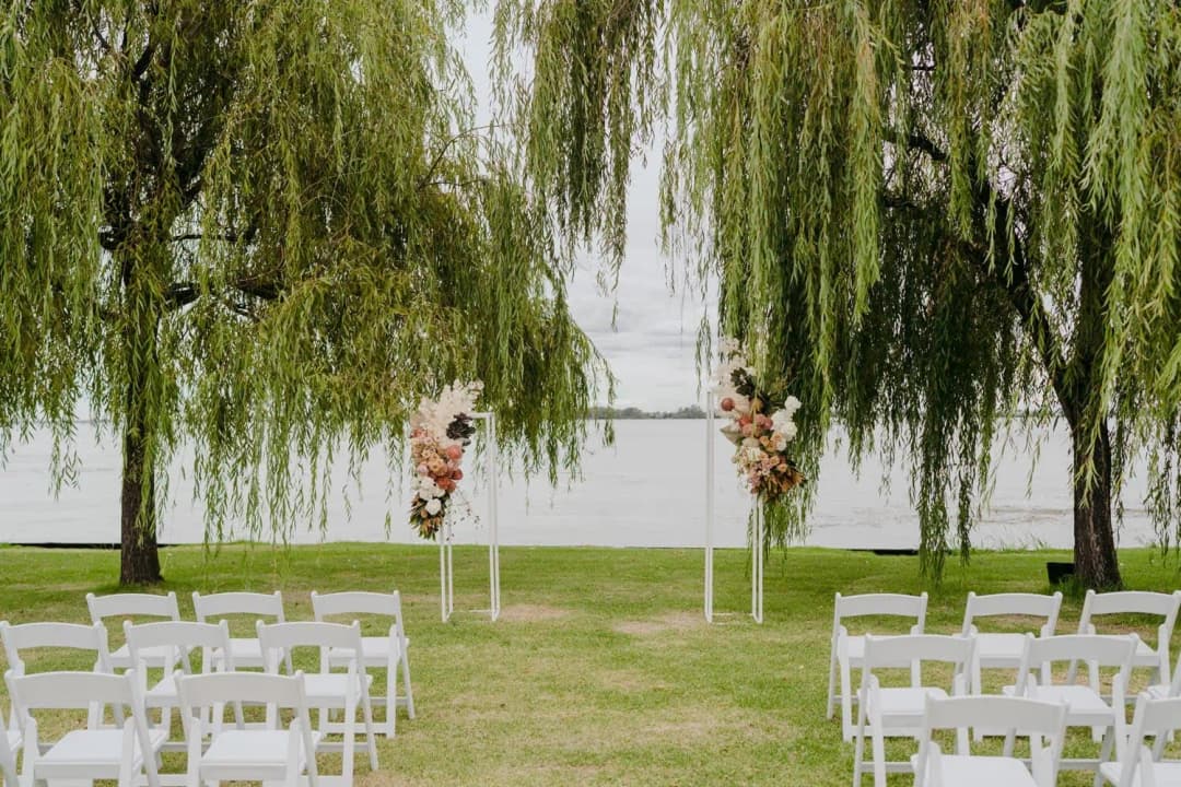 White ceremony chairs facing floral stands beneath willow trees beside a lake