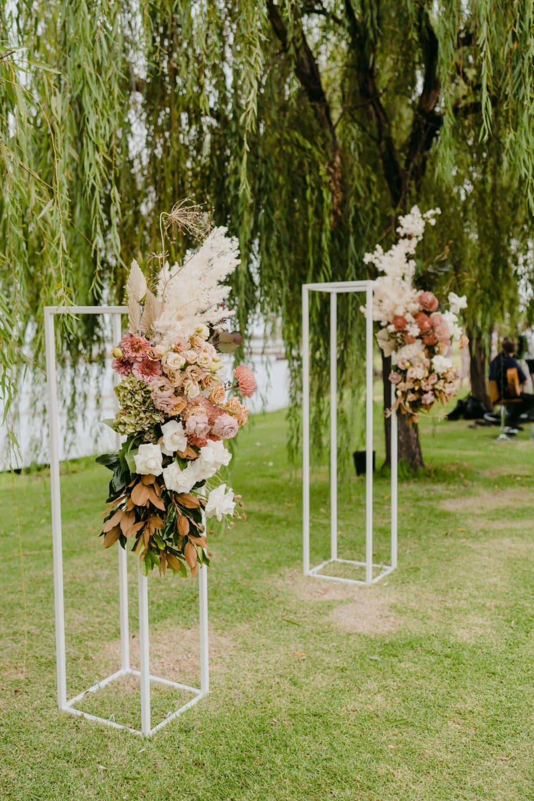 Peach and white flowers arranged on tall white ceremony stands beneath willow branches